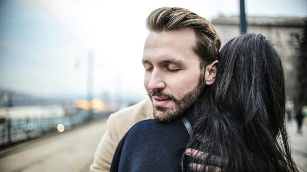A man with his eyes closed hugs a person with long dark hair outdoors.