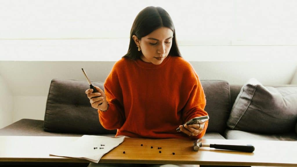 A woman assembling small hardware pieces while checking her phone at a table.