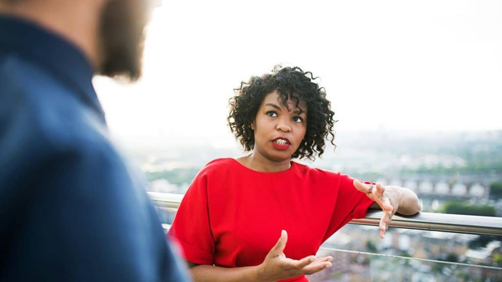 A woman in a red top gesturing while speaking to someone outdoors.