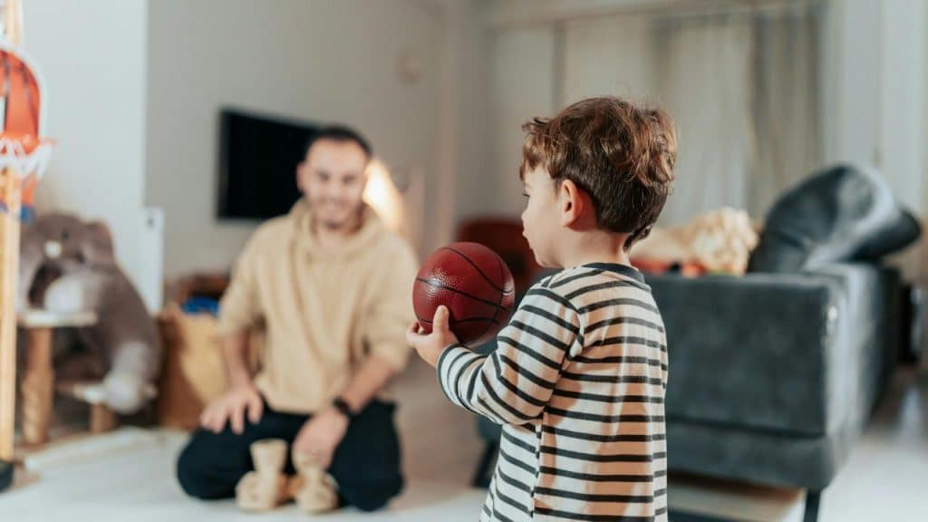 A young child holding a basketball while an adult watches in a living room