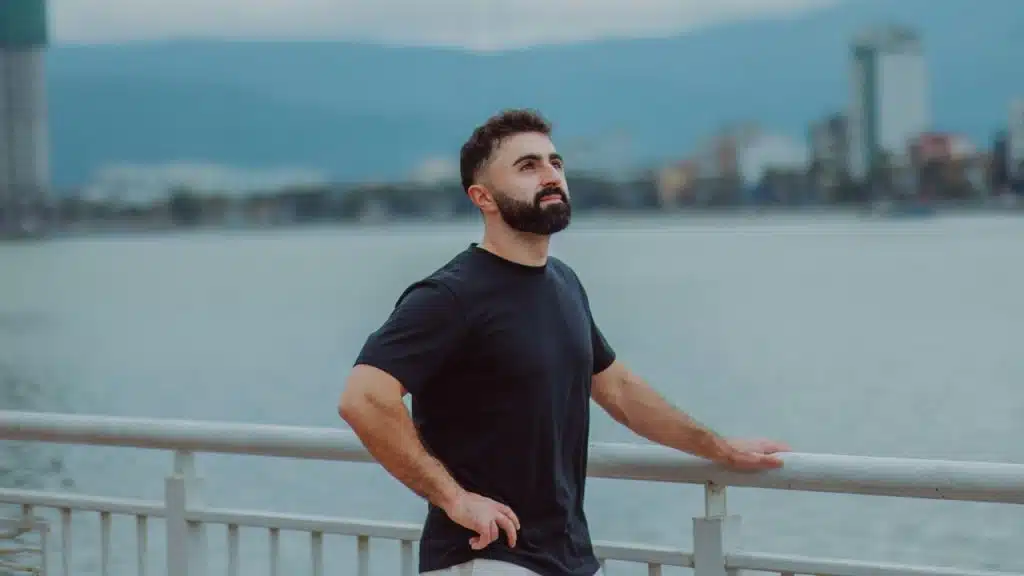 A man in a black t-shirt leans against a railing overlooking a body of water.