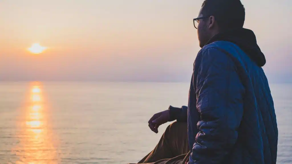 A man sits by the water watching the sun set over the horizon.