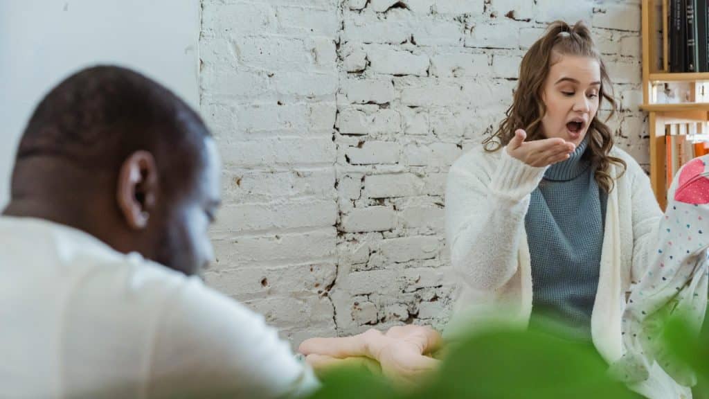 A woman in a white cardigan gestures while talking to a man in a room.