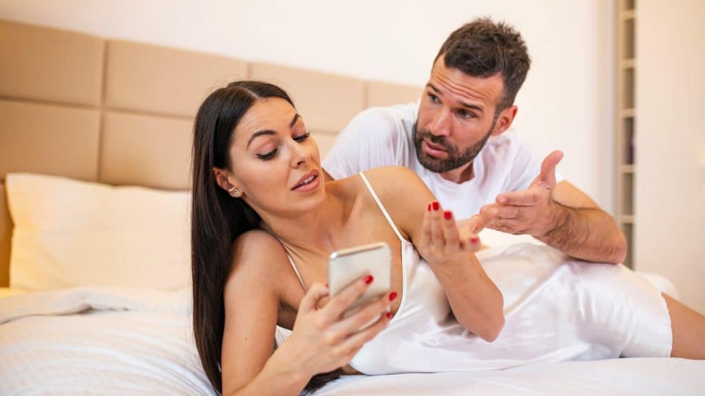 A woman lying on a bed looks at a phone while a man gestures beside her.