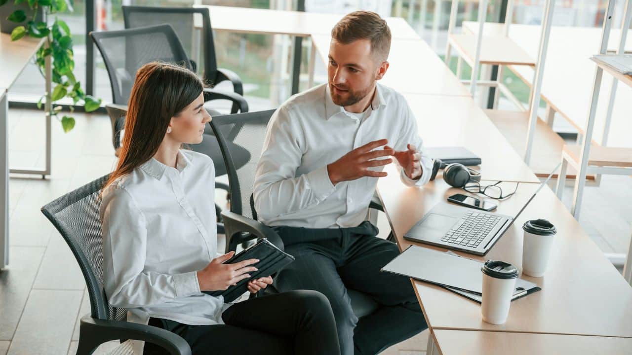 A man gestures while speaking to a woman at a long desk in a bright office.