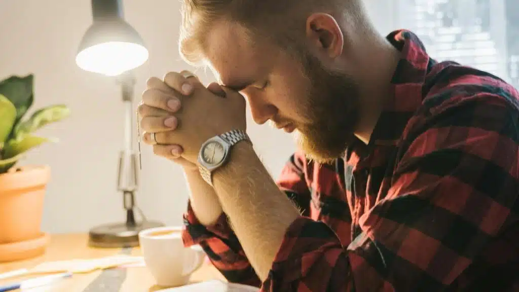 A bearded man praying at a desk with a cup of coffee and a lamp nearby.