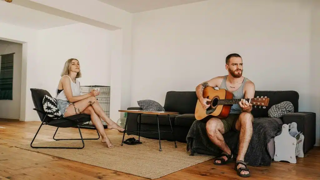 A man plays an acoustic guitar on a couch while a woman watches from a chair.