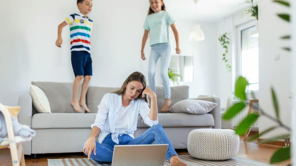 A woman sits on the floor with a laptop while two children jump on a sofa.