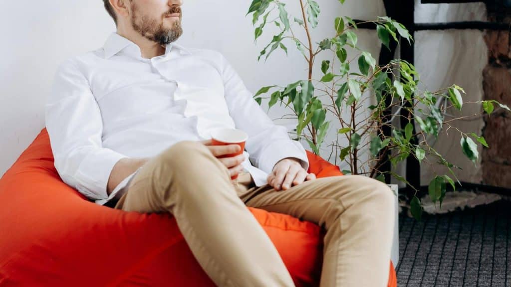 A bearded man in a white shirt sitting on a red beanbag chair holding a cup indoors.