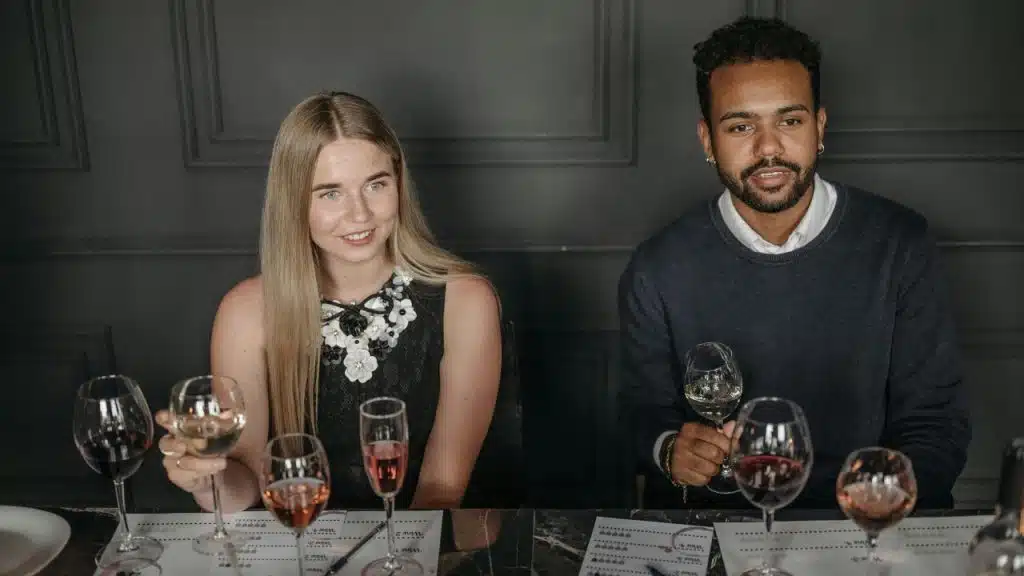 A man and woman sit at a table with wine glasses, looking ahead.