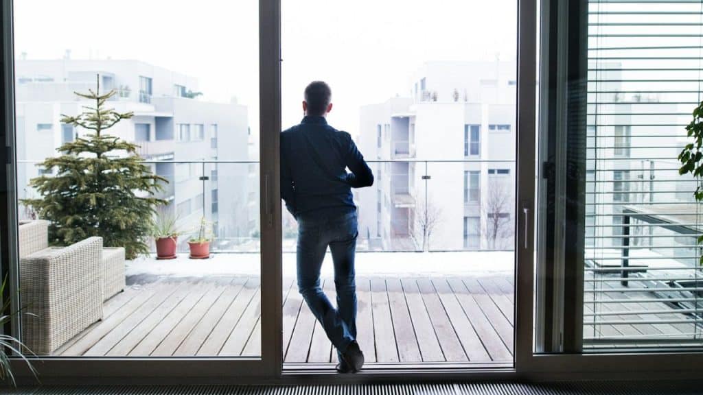 A man stands on a wooden balcony looking out at several white apartment buildings.