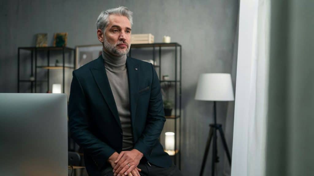 A man with grey hair wears a dark blazer and looks out of a window indoors.