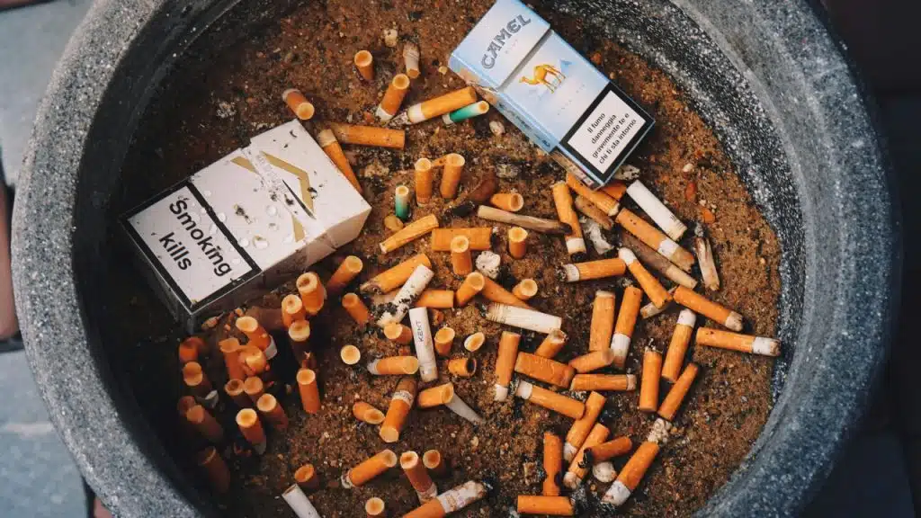 An overhead view of an ashtray filled with sand, many cigarette butts, and two empty packs.