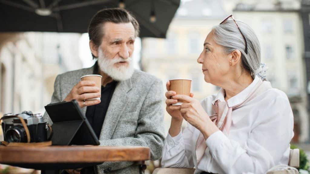 An older man and woman sit at an outdoor table holding cups and looking at each other.