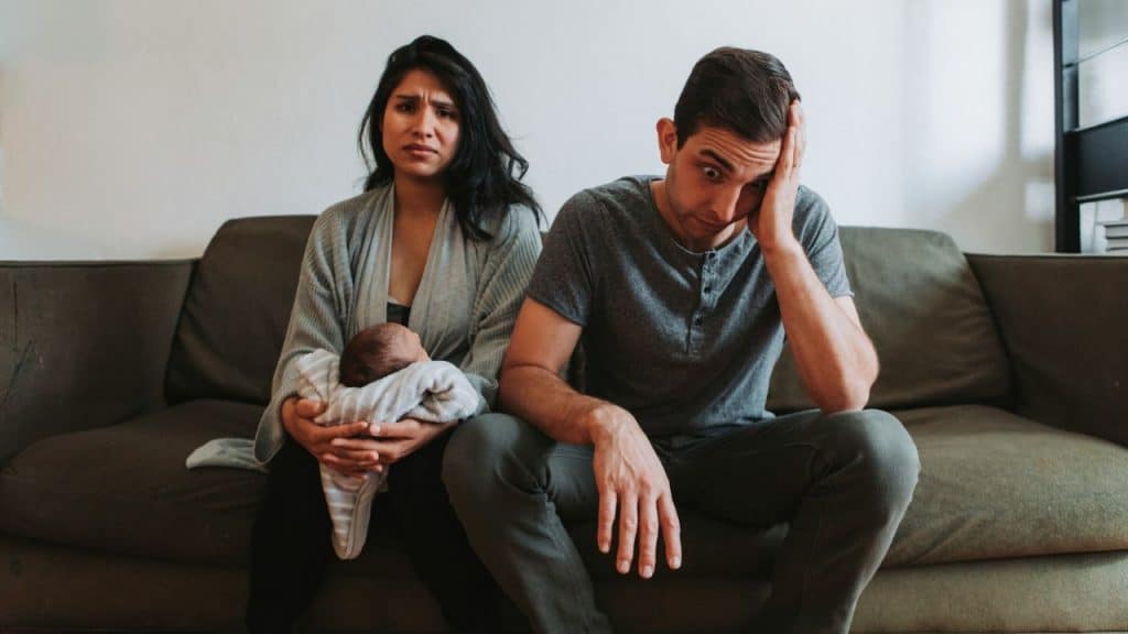 A woman holding a baby and a man with his hand on his head sit together.