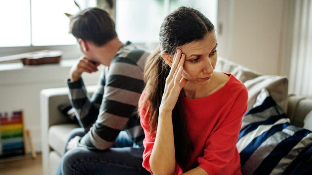 A man and woman sit on a couch, facing away from each other, appearing distressed.