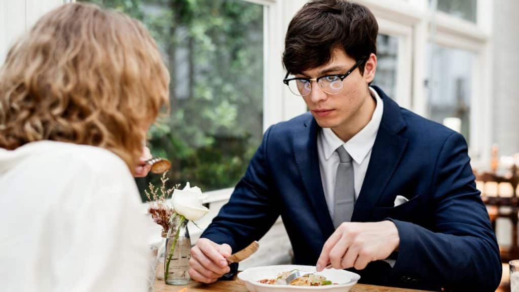 A man in a suit and glasses eats dinner across from a person with curly hair.
