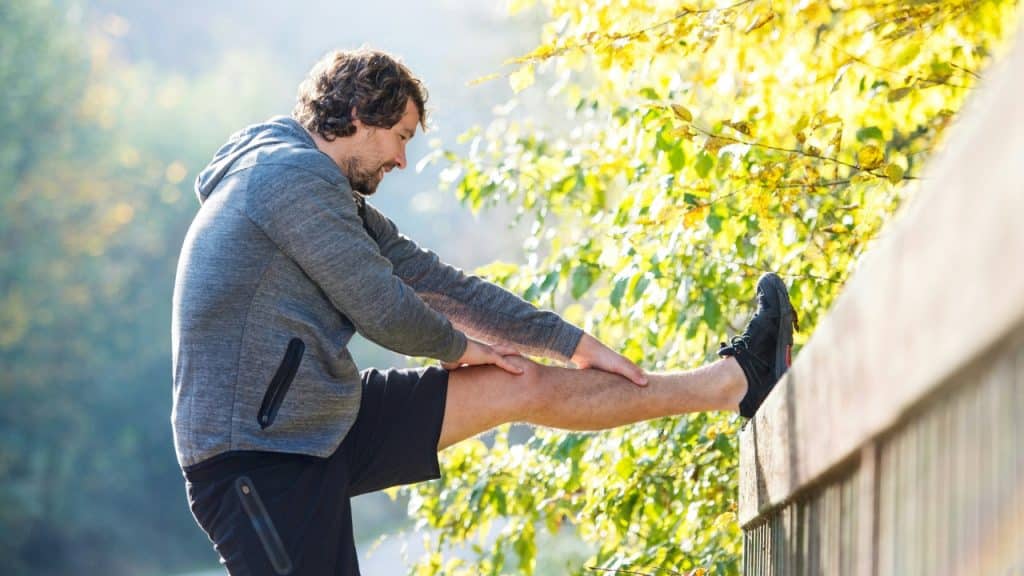 A man in athletic wear stretches his leg on a wooden railing outdoors.