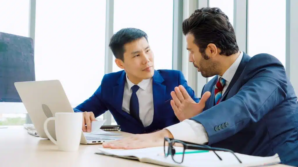 Two professional men sit at a desk while one gestures during a serious conversation.