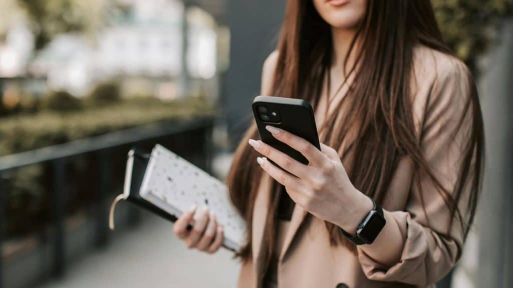 A woman outdoors looking at her smartphone while holding a notebook.
