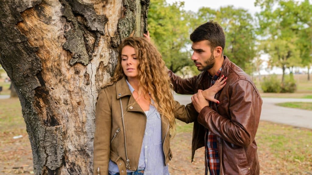 A man in a leather jacket holds a woman's arm near a large tree trunk.