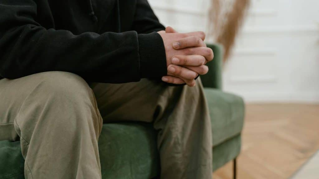 A person sitting on a couch with hands clasped, appearing thoughtful.