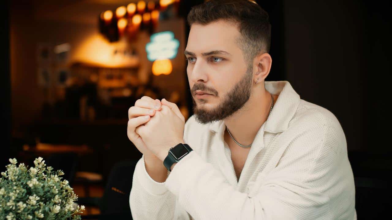 A bearded man in a light sweater sitting thoughtfully at a table