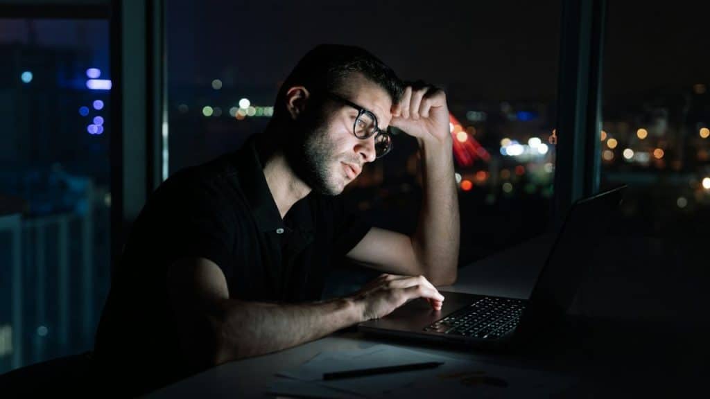 A man wearing glasses looks at a laptop screen in a dark room with city lights.