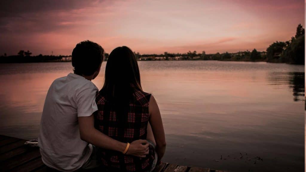 A man and a woman are watching the sunset while sitting on a rock near a body of water.