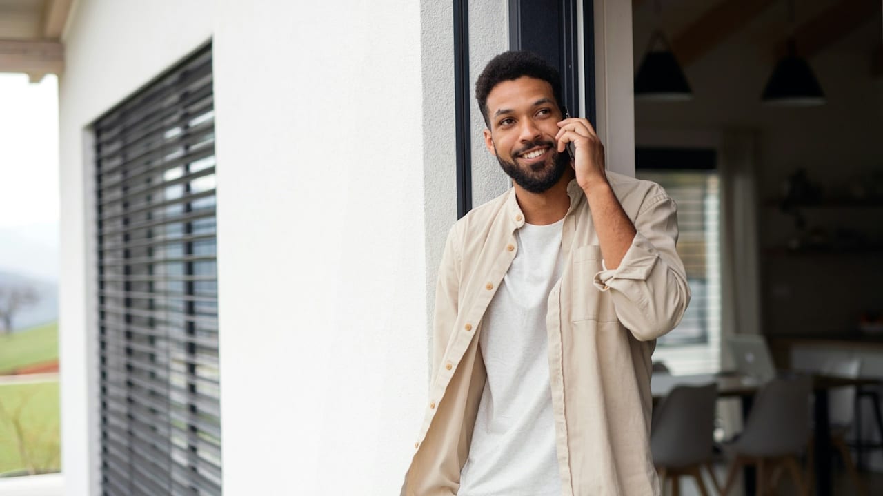 A bearded smiling man is standing in the doorway of his home and talking on the phone.