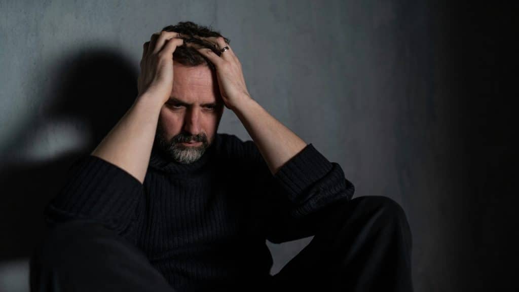 A man with a beard sits against a dark wall with both hands clutching his hair.