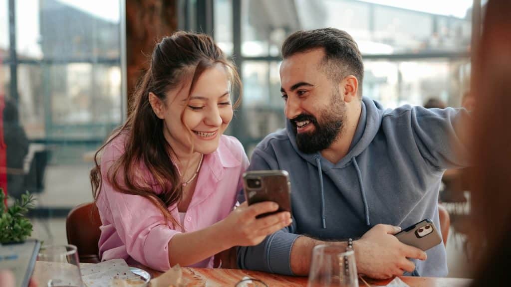 A woman and a man smile while looking at a smartphone together at table.