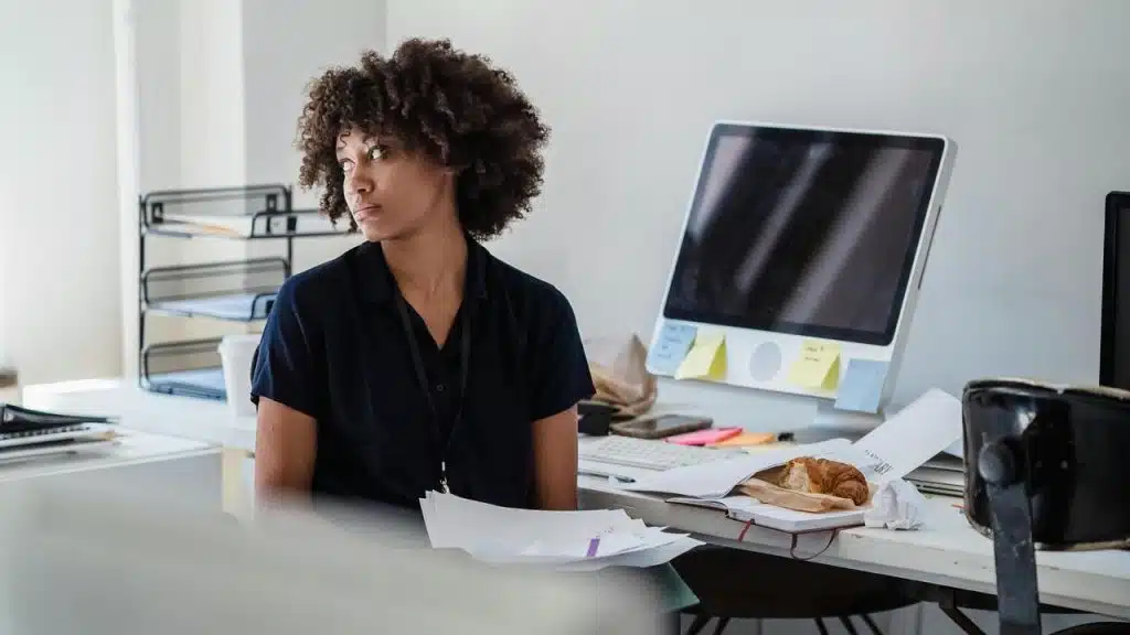 A woman sitting at a cluttered desk in an office, looking away thoughtfully.