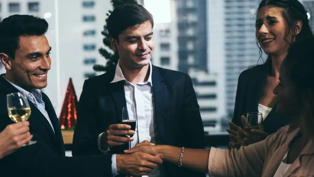 People in business attire hold glasses and shake hands in a city office setting.