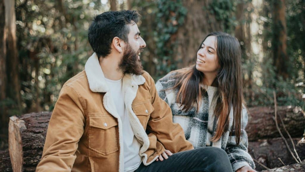 A man with a beard and a woman sit together, both smiling and looking at each other.