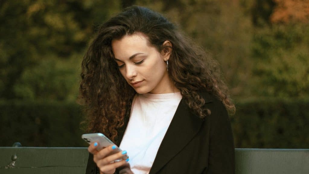 A woman with long curly hair looks down while holding a mobile phone outdoors.