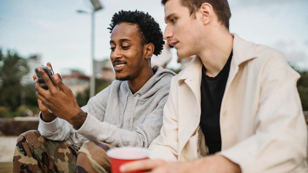 A smiling man showing something on his phone to a friend while sitting outdoors.