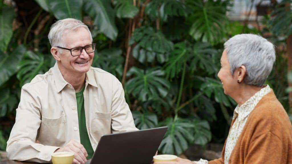 An older man and woman sit at a table outdoors with a laptop and coffee.