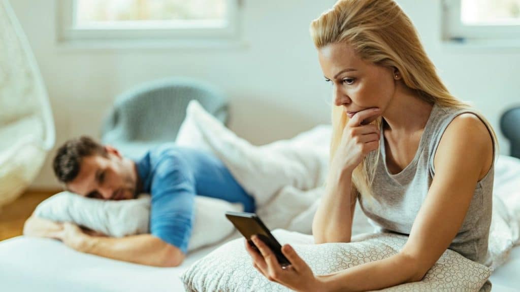 A woman looks at a smartphone on a bed while a man sleeps behind her.
