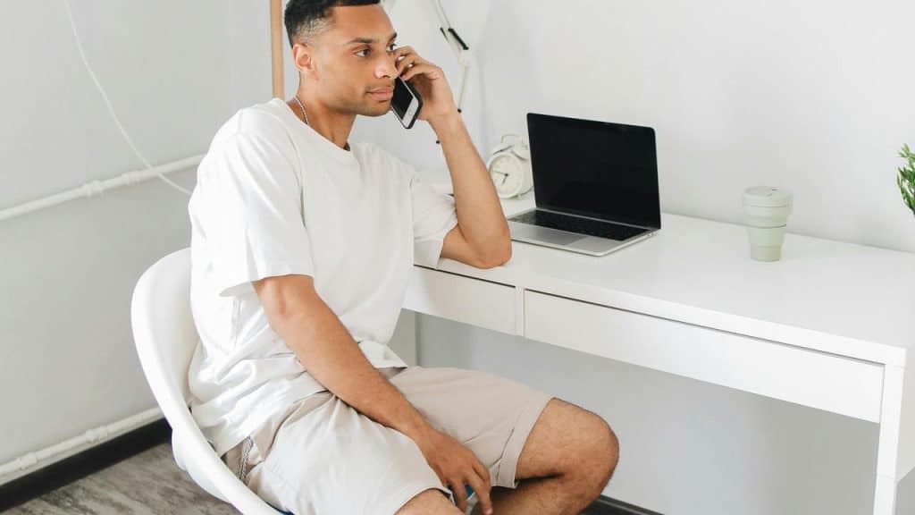 A man sitting at a desk talking on a smartphone with a laptop nearby.
