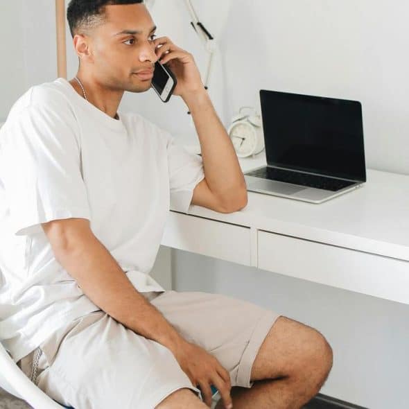 A man sitting at a desk talking on a smartphone with a laptop nearby.