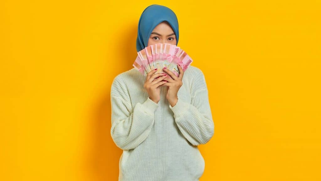 Woman in a blue headscarf holding pink banknotes in front of her face against a yellow background.