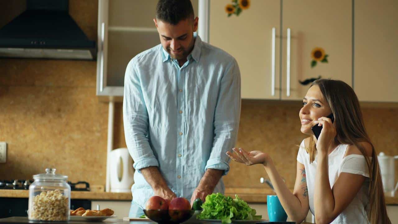 A man cutting salad while her wife is talking on phone.