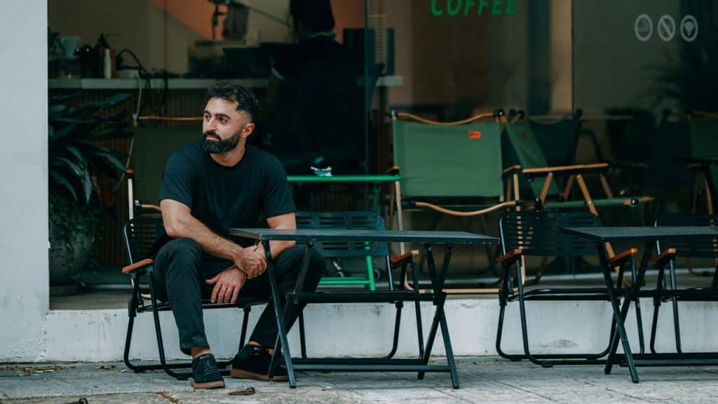 A bearded man in a black outfit sitting at an outdoor café table, looking to the side.