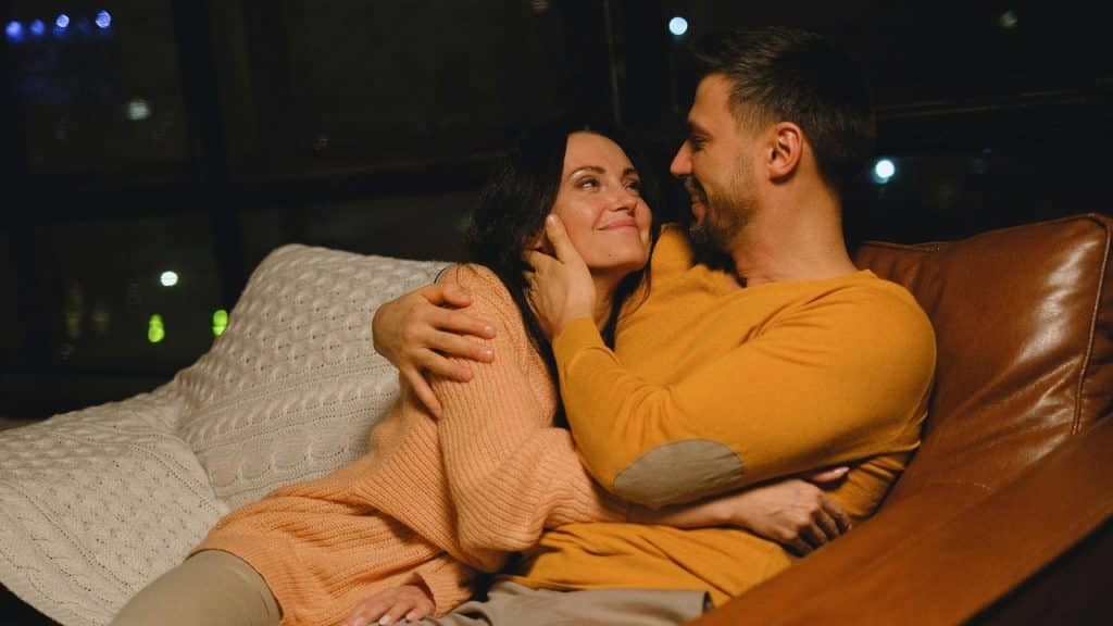A man and woman sitting together on a brown leather sofa in a dimly lit room.