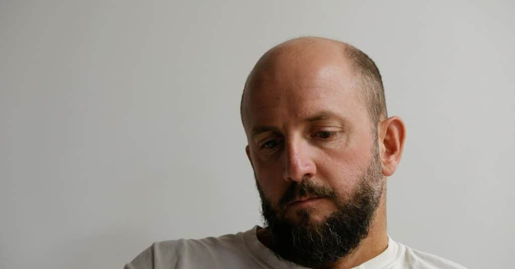 A bearded man looking down with a thoughtful expression against a plain background.
