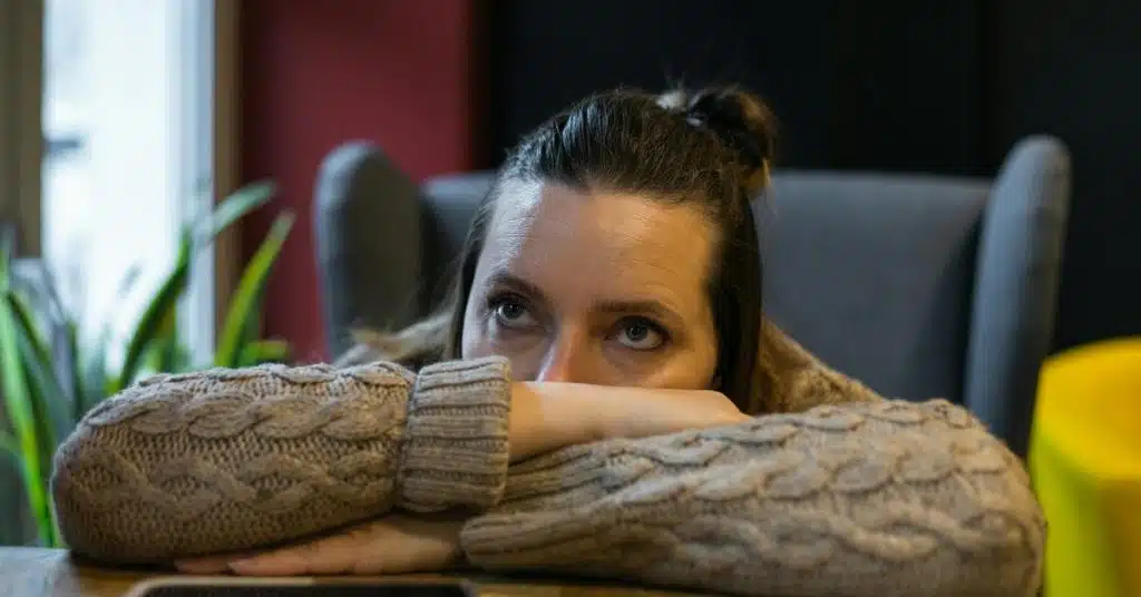 A woman resting her head on folded arms, gazing upward with a pensive expression indoors.
