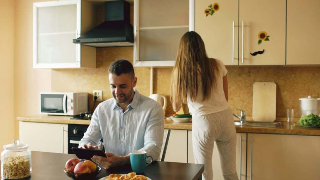 A man uses a tablet at a table while a woman stands at a kitchen counter.