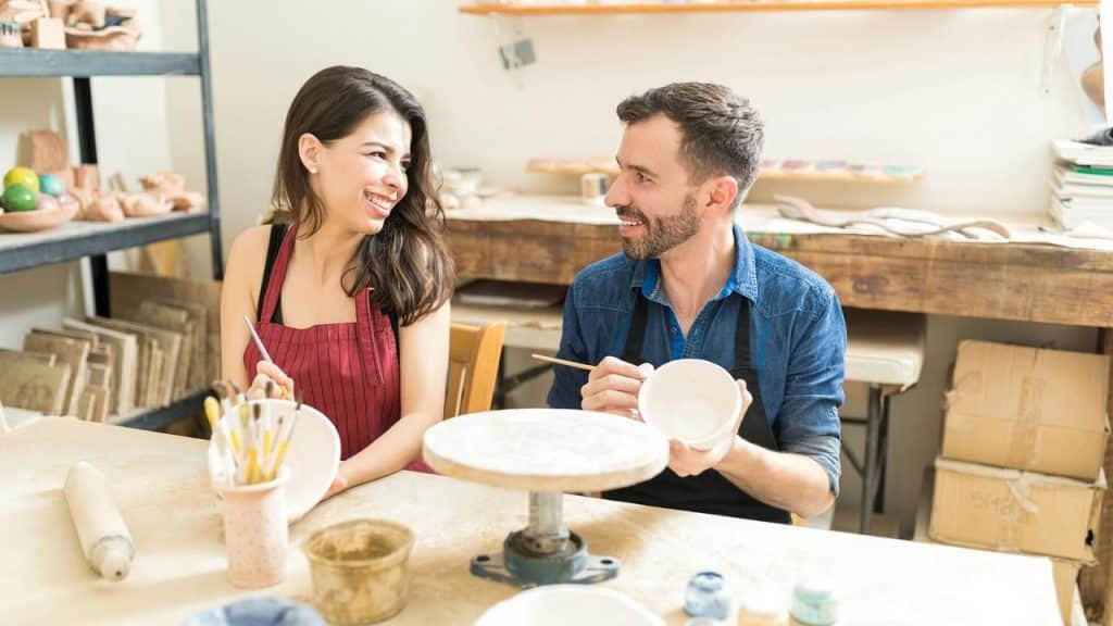 A couple smiles at each other while painting pottery in a ceramics studio.
