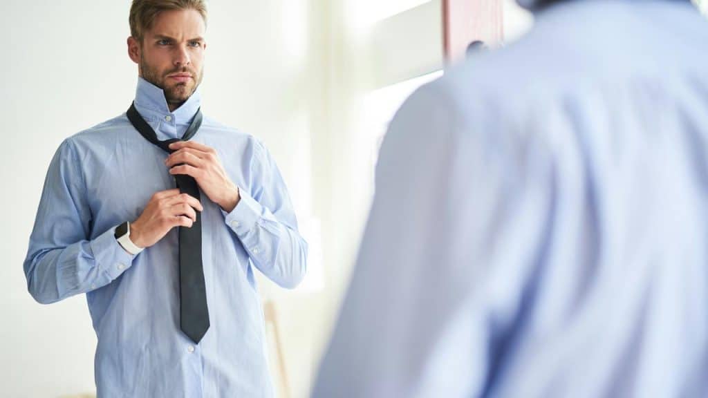 A man in a blue shirt tying a black tie in front of a mirror.